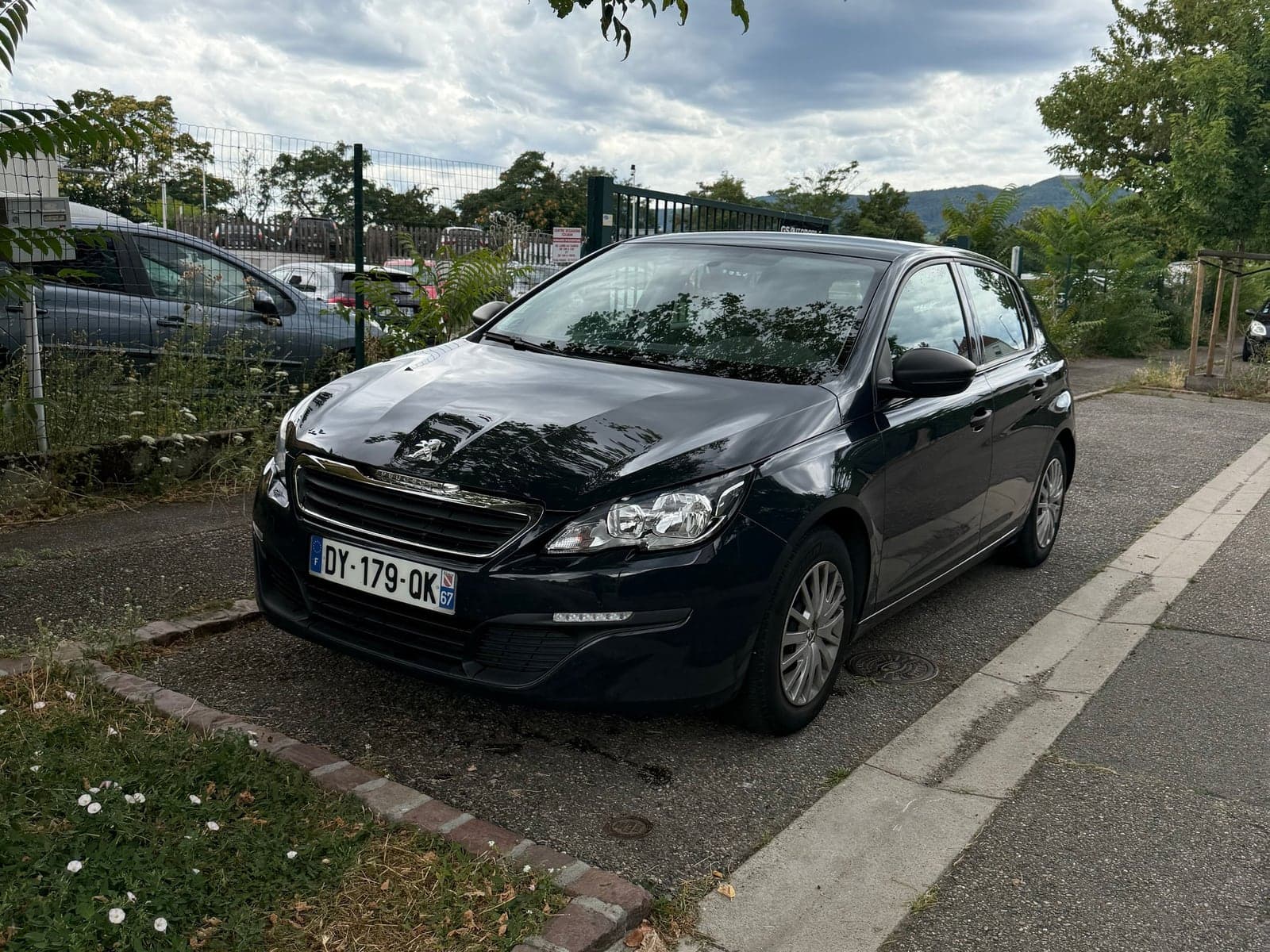 Peugeot 308 noire photographiée au téléphone sur un parking, avant traitement : ciel gris, plaque lisible